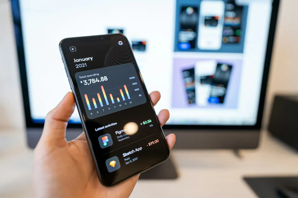 A hand holds a phone showing a fitness dashboard beside a monitor.